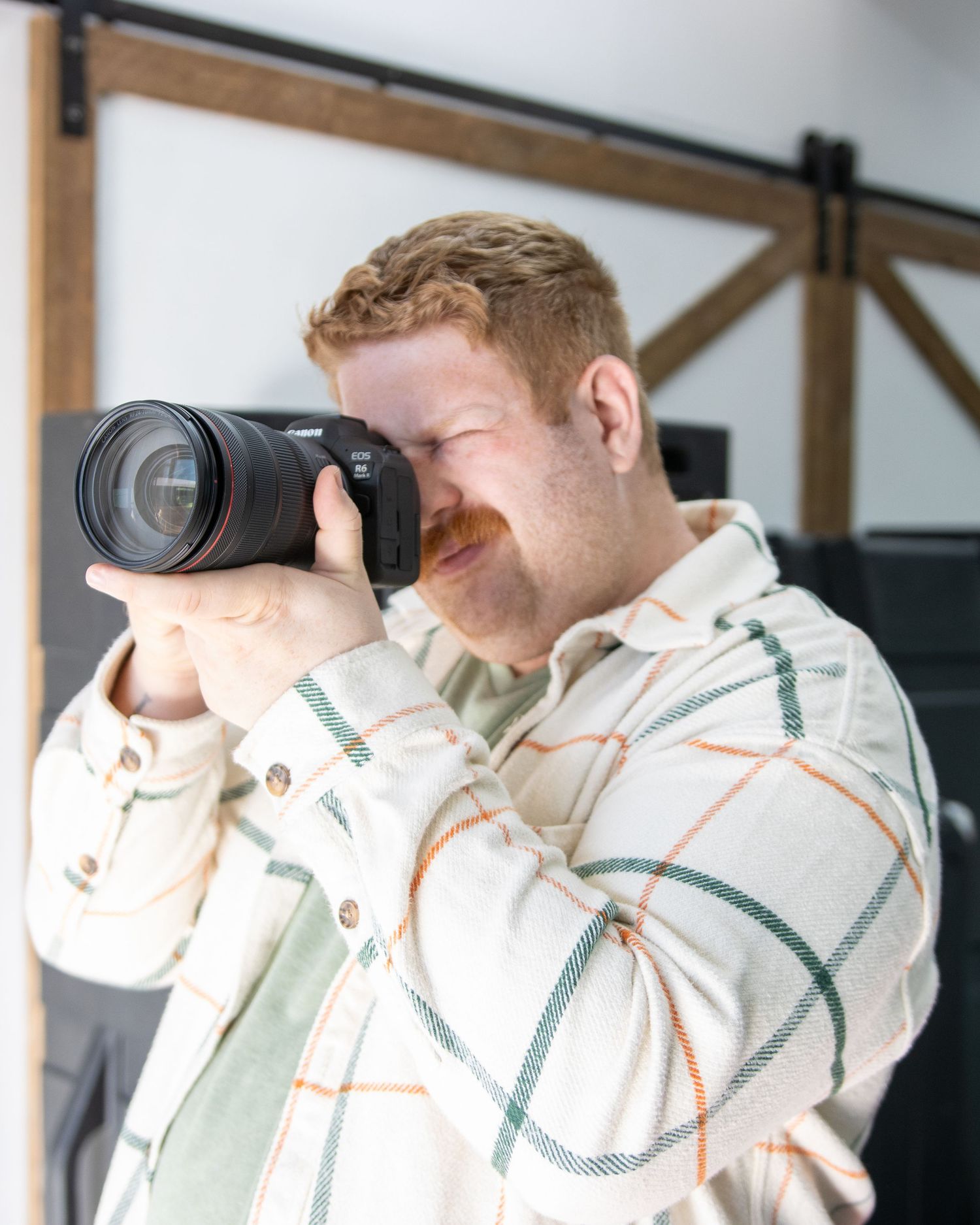 A person wearing a white plaid shirt looks through a long telephoto lens on a professional camera with dark background.