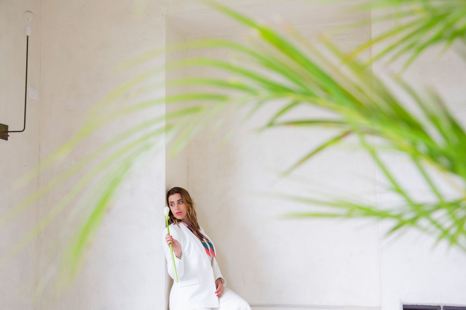 Person in white attire sits against white wall with palm leaf shadows creating artistic tropical ambiance.