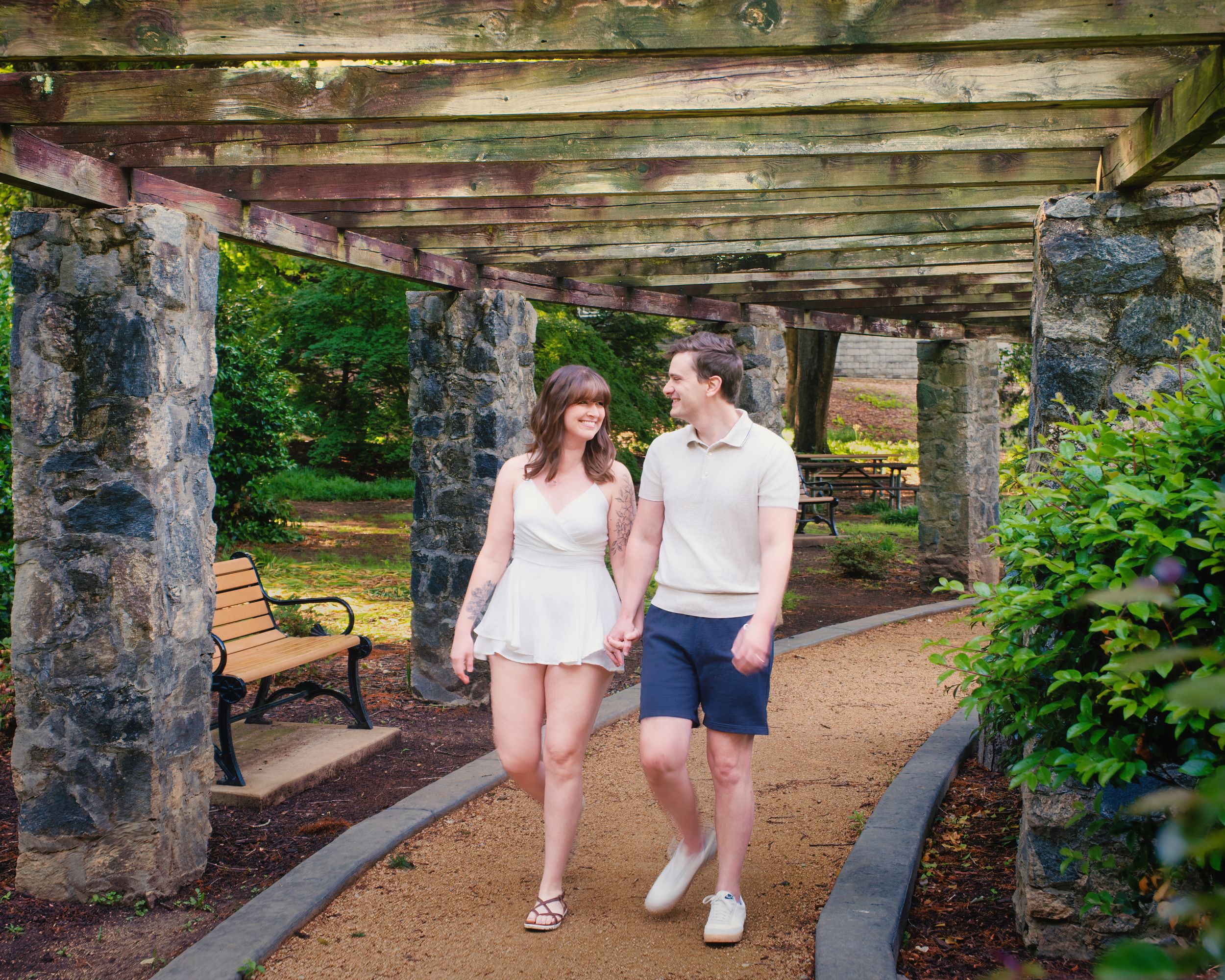 A couple walks together under a stone pergola surrounded by lush gardens on a summer day.