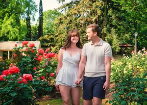 A couple strolls through a vibrant garden path lined with blooming red roses and lush greenery on a sunny day.