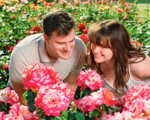 Couple smiling together while leaning over vibrant pink blooms in a colorful garden filled with roses.
