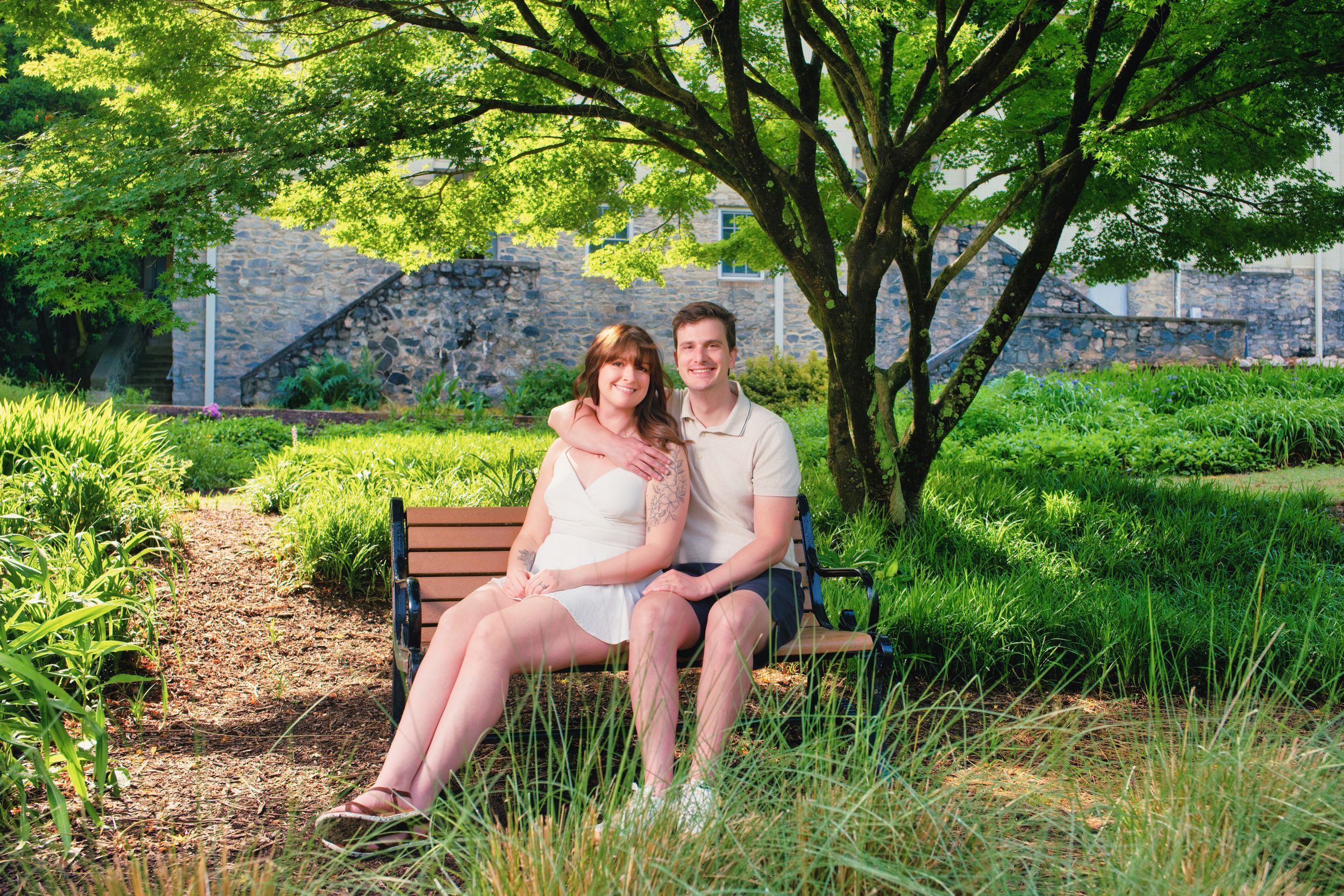 A couple sitting together on a wooden bench in a serene garden with trees and stone walls in the background.
