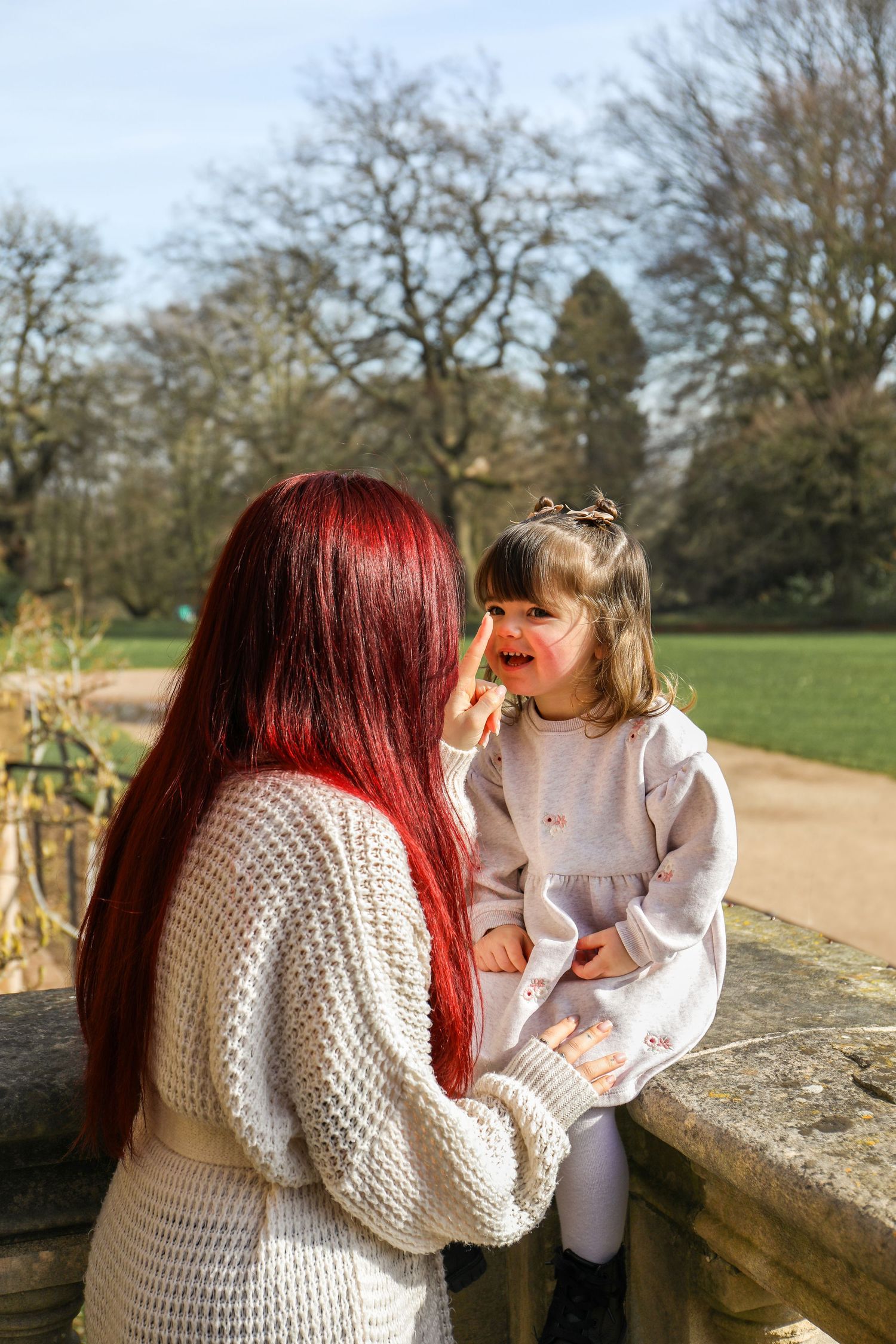 Two people share a sweet moment while sitting on a stone wall in a park on a sunny day, during Family shoot in Nottingham