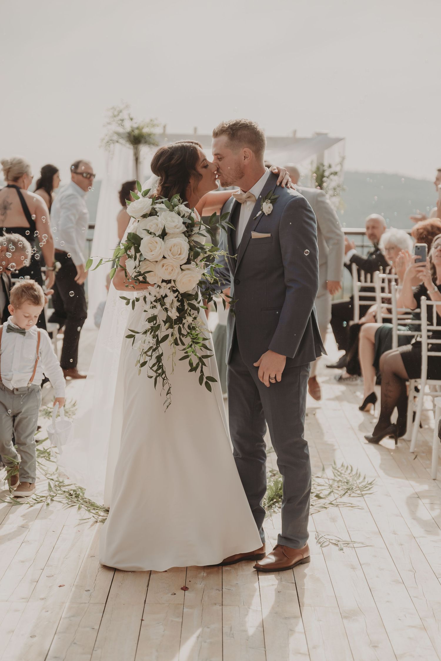 Elegant wedding ceremony kiss on wooden deck with white floral arrangements.