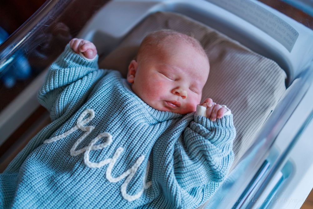 Sleeping newborn baby wearing light blue knit sweater in bassinet.