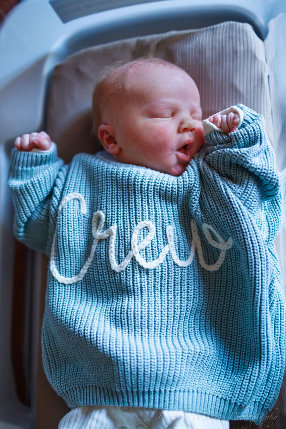 Baby wearing blue knit sweater with white lettering sleeping in bassinet.