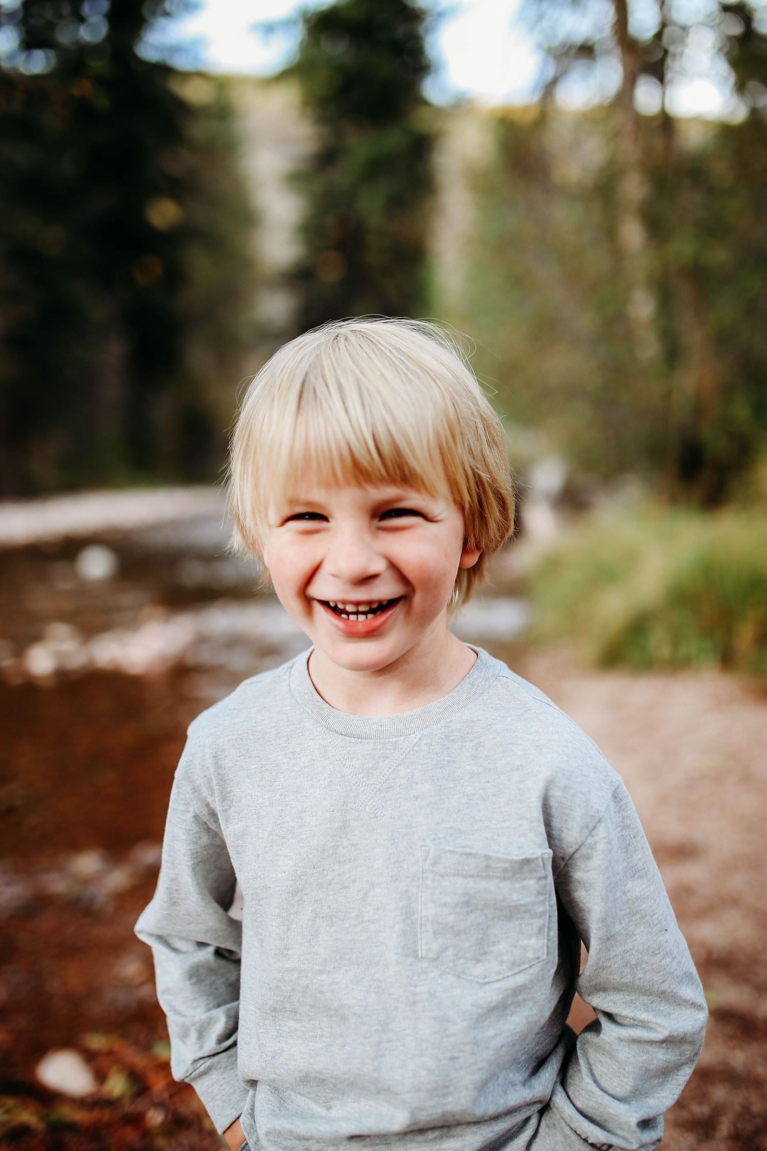 A small child in a grey sweatshirt smiles joyfully while standing outdoors near a stream.