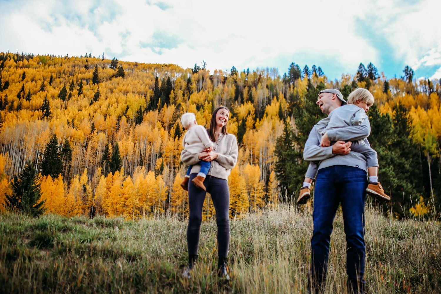 Two couples holding young children stand in a grassy meadow with golden autumn aspens on the mountainside behind them.
