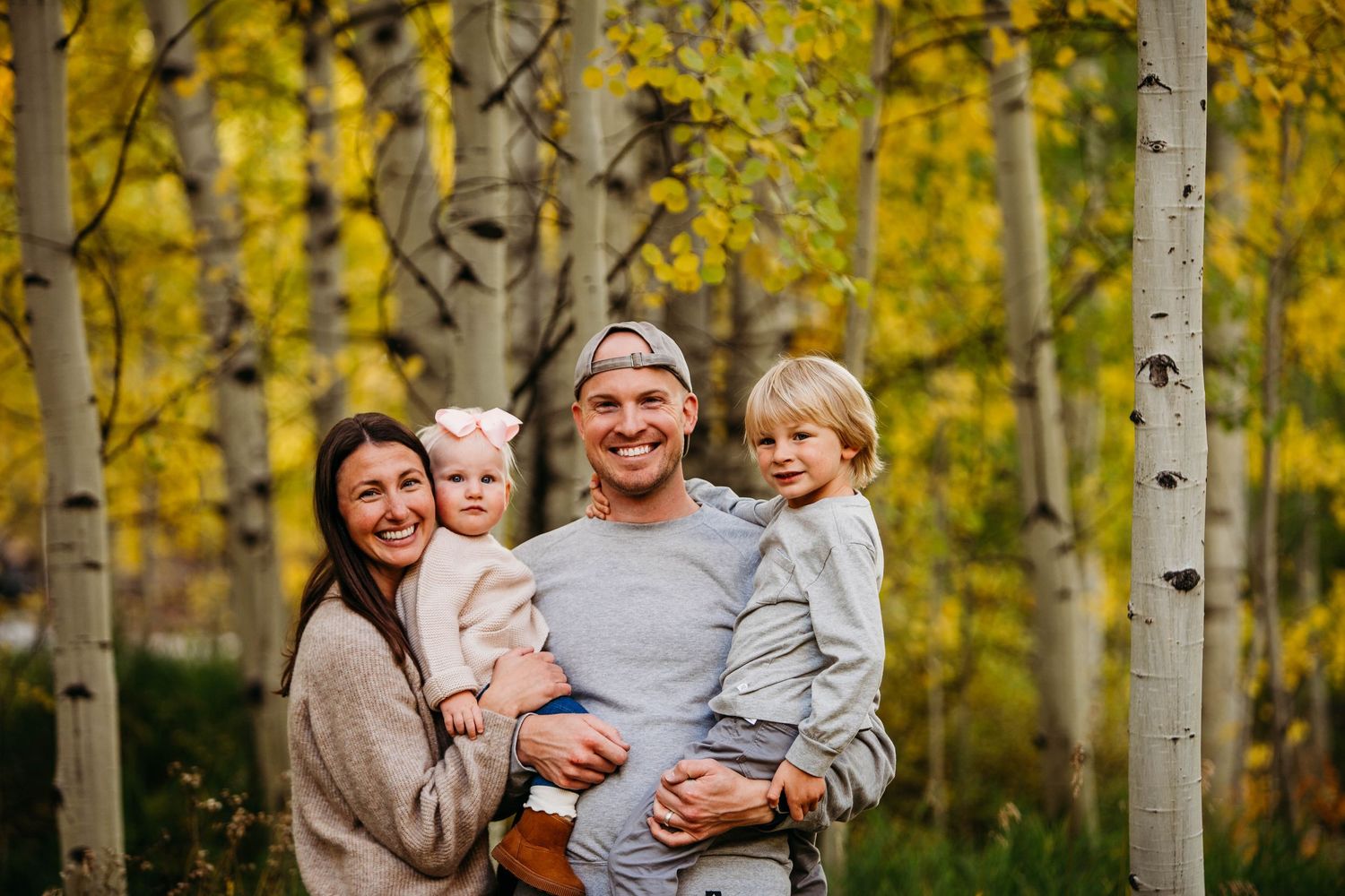 A family stands together smiling surrounded by aspen trees with golden autumn foliage in a forest.