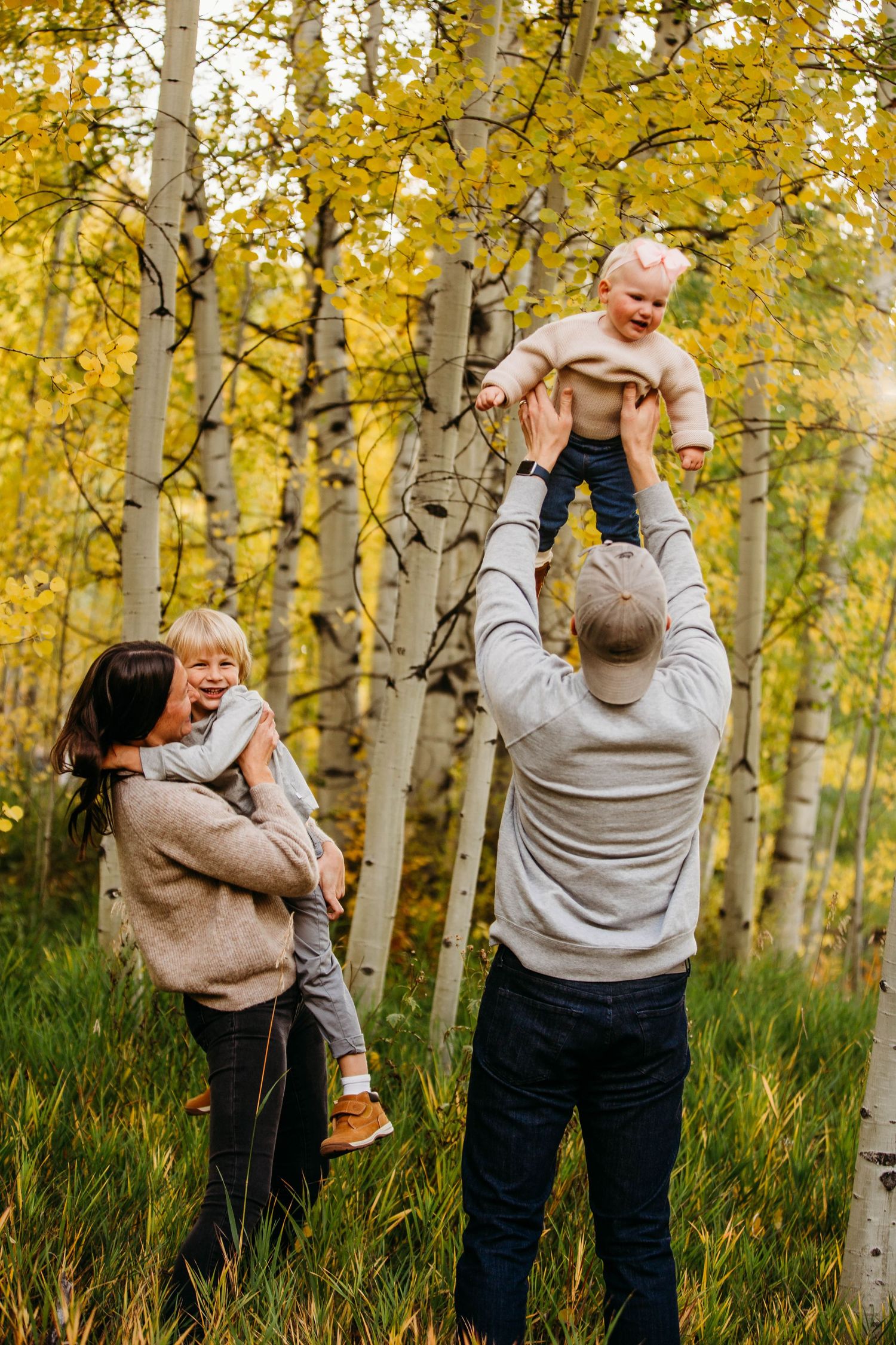 Joyful outdoor portrait in autumn aspen grove with family playing and lifting children against golden foliage.