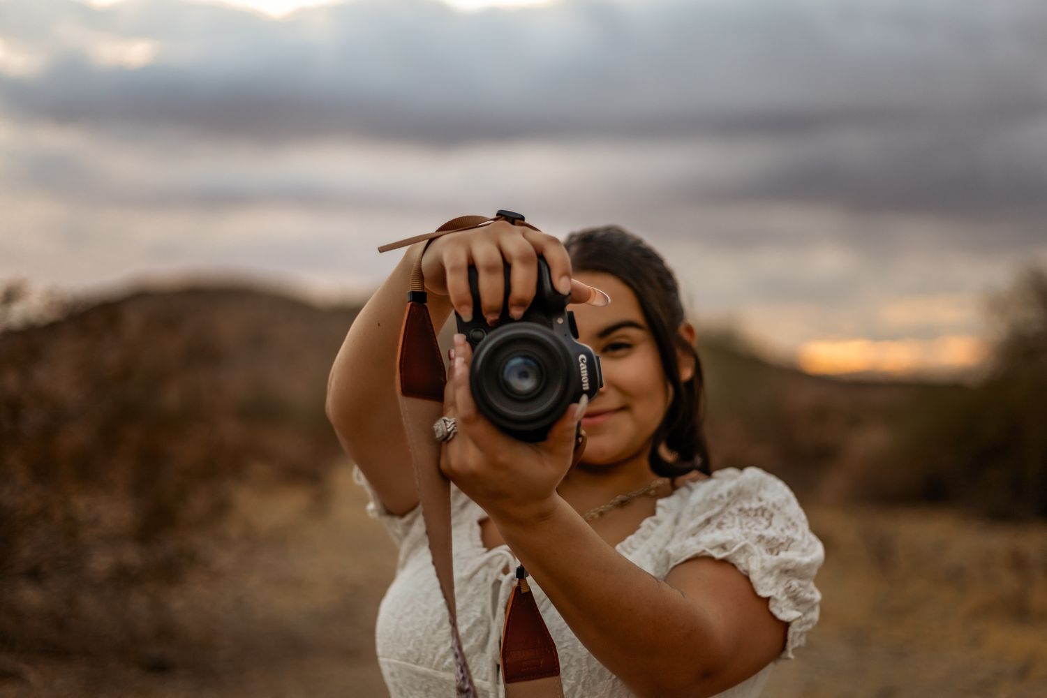 A photographer wearing a white lace dress takes a photo with a digital camera against a sunset desert backdrop.