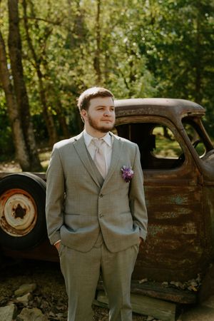 Groom in gray suit poses next to vintage rustic truck in wooded setting.