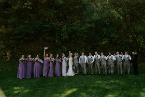 Wedding party in purple dresses and gray suits standing in formation against green foliage.
