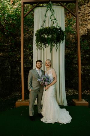 A wedding couple poses under an elegant pergola decorated with greenery and white drapes.
