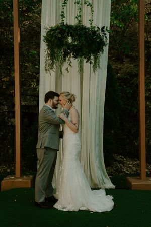 A tender wedding moment captured beneath a draped pergola with hanging greenery.