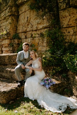 Newlyweds share a relaxed moment while seated against a historic stone wall backdrop.
