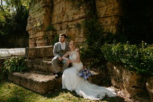 Wedding couple poses casually by a weathered stone wall surrounded by garden foliage.