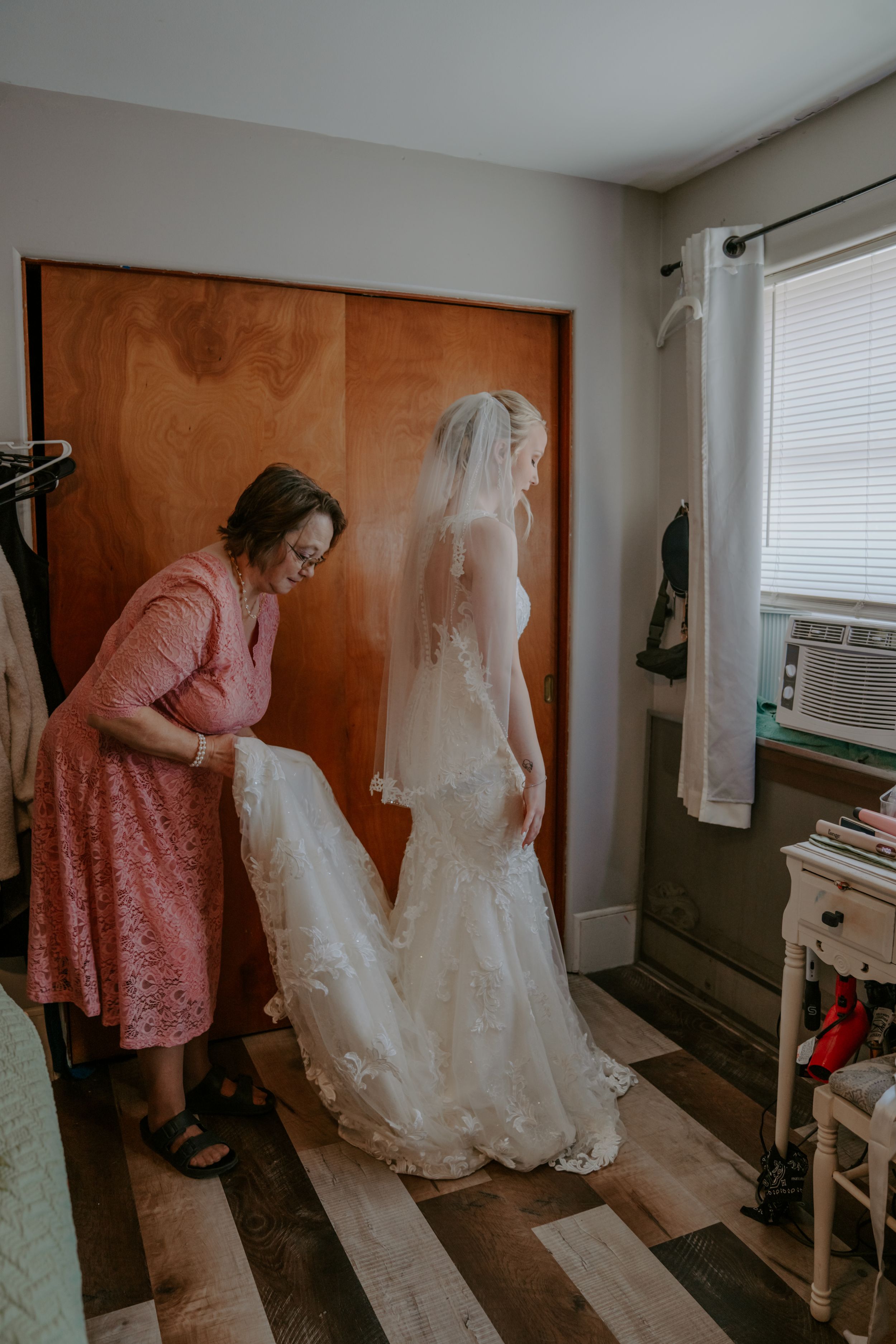 Wedding dress preparations in a sunlit room with pink bridesmaid dresses visible.