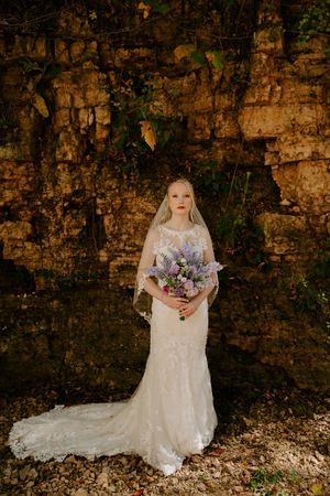 Bride in lace gown poses against rustic stone wall holding purple bouquet.