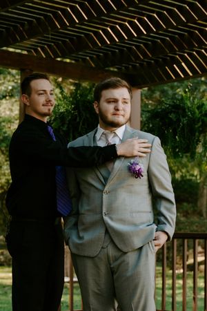 Groomsmen adjusting suit and boutonniere under wooden pergola.