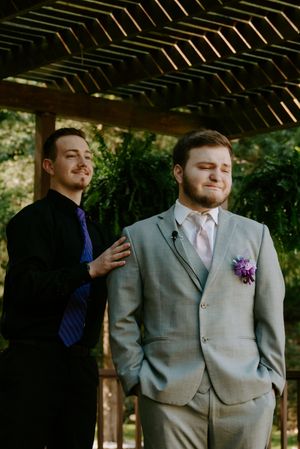 Groom in light gray suit with purple boutonniere stands under wooden pergola.