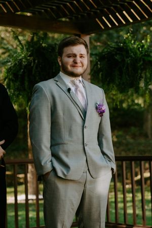A groom in a light gray suit with a purple boutonniere poses outdoors during a wedding ceremony.