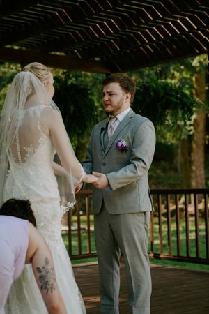 Wedding ceremony details showing lace dress and gray suit under a wooden pergola.