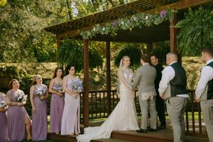 Wedding party in purple dresses and gray suits gathered under trees for outdoor ceremony.