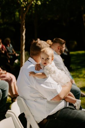 A small child in white formal wear being held during an outdoor wedding ceremony.