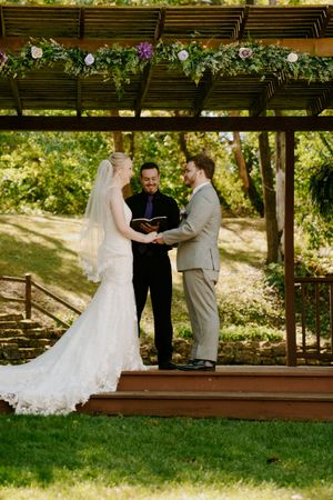 A wedding ceremony takes place under a rustic wooden pergola decorated with flowers in a garden setting.