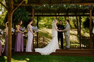 Wedding party stands in line under a wooden pergola with purple and white floral decorations.