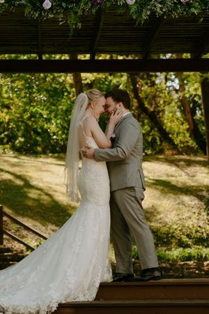 Newlyweds share their first kiss as a married couple under a wooden pergola in a garden setting.