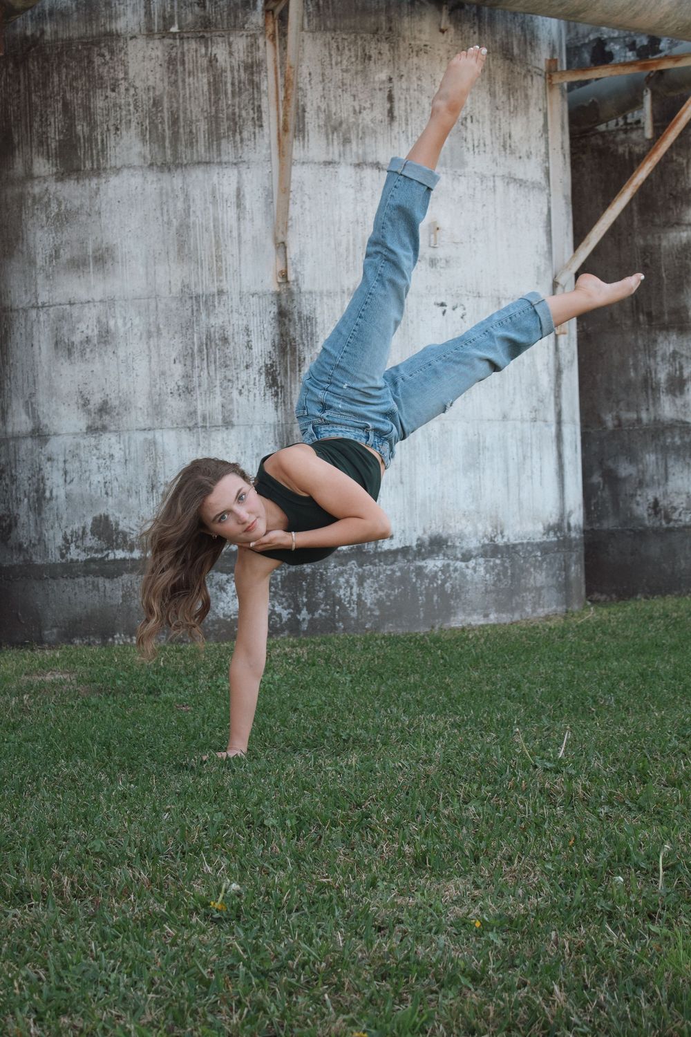 Young dancer performs a dramatic one-armed handstand on grass against a concrete wall.