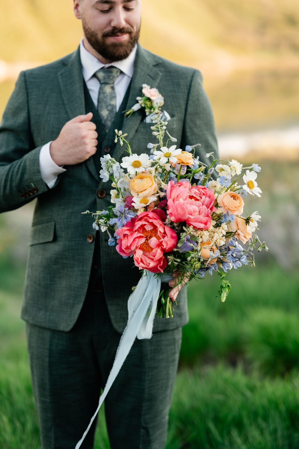 Colorful spring wedding bouquet with coral peonies and daisies held against a green suit jacket.