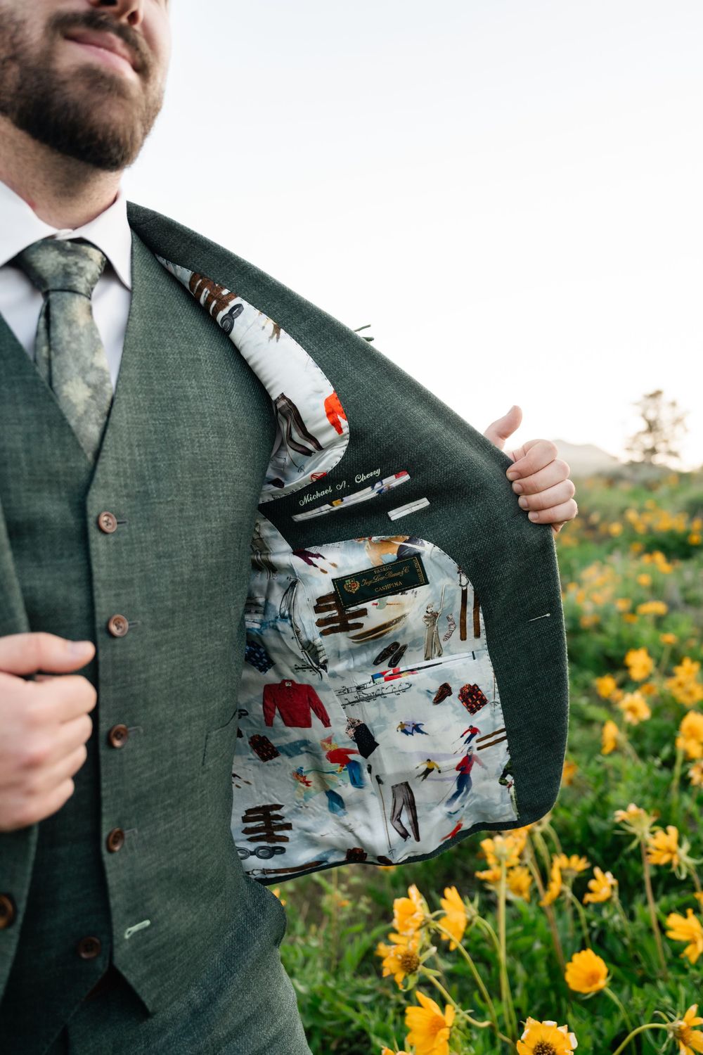 A groom in a green tweed vest shows off his custom-printed suit lining while standing in a field of yellow flowers.