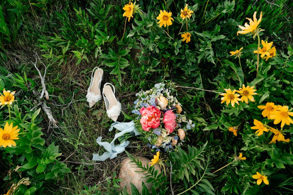 White bridal shoes and pink floral bouquet arranged among yellow daisies in a garden setting.