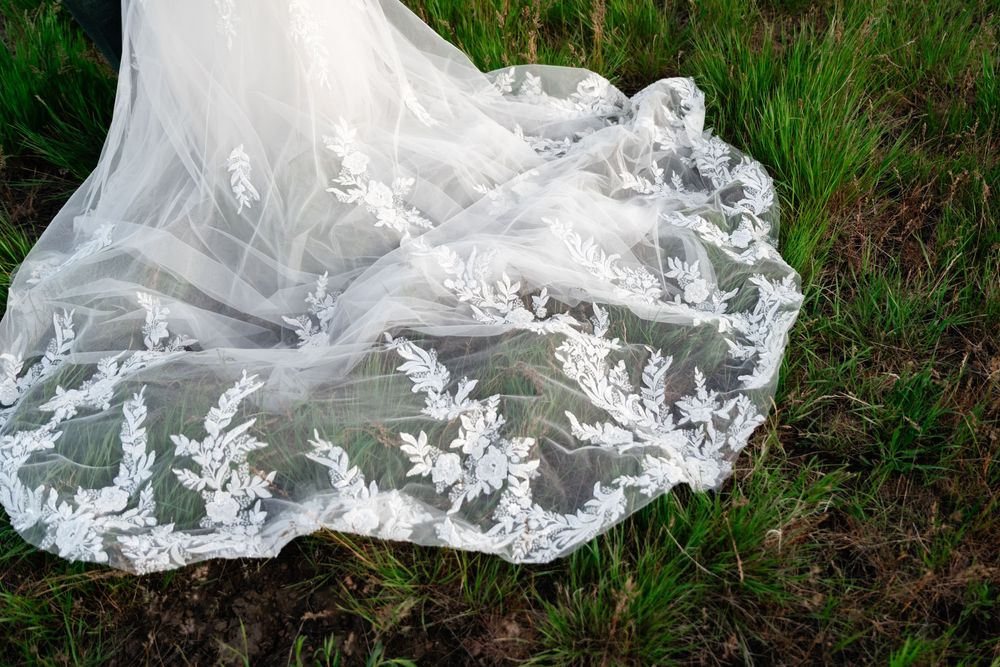 Bottom hem of an ornate white wedding dress trailing through green grass.
