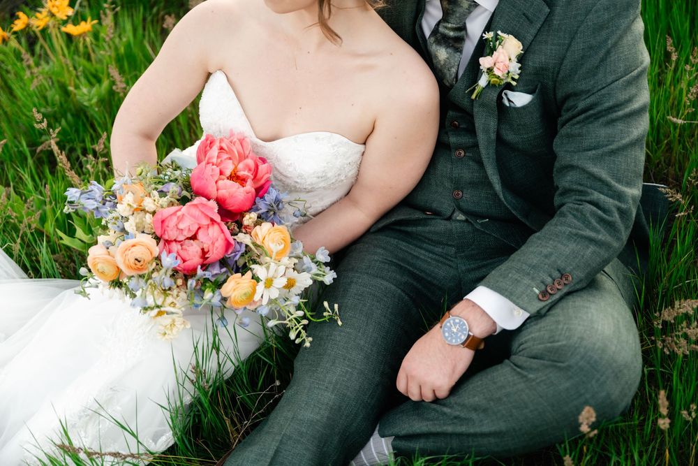 Wedding couple sitting together in a grassy meadow holding vibrant coral and orange floral bouquets.