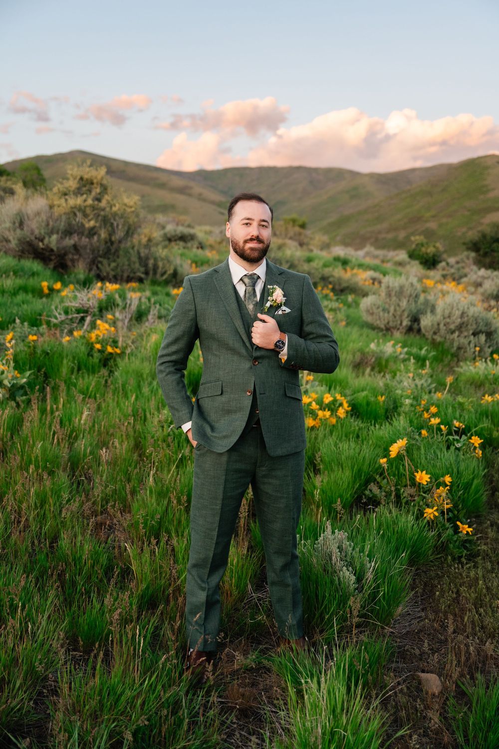 Groom in dark suit poses in field of yellow wildflowers with mountains in background at sunset.