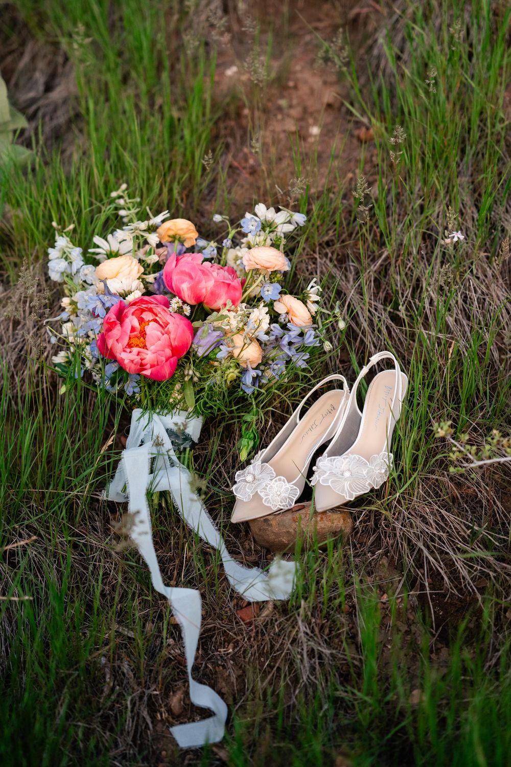 Wedding details flat lay showing bridal shoes, delicate lace pieces and a coral peony bouquet in grass.