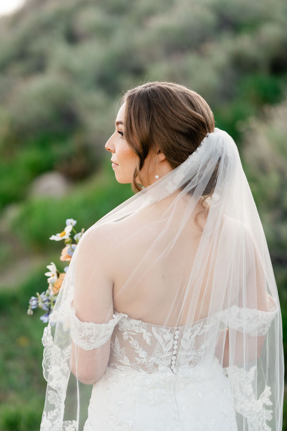 A bride in a white lace off-shoulder wedding dress and long veil stands against a natural outdoor background.