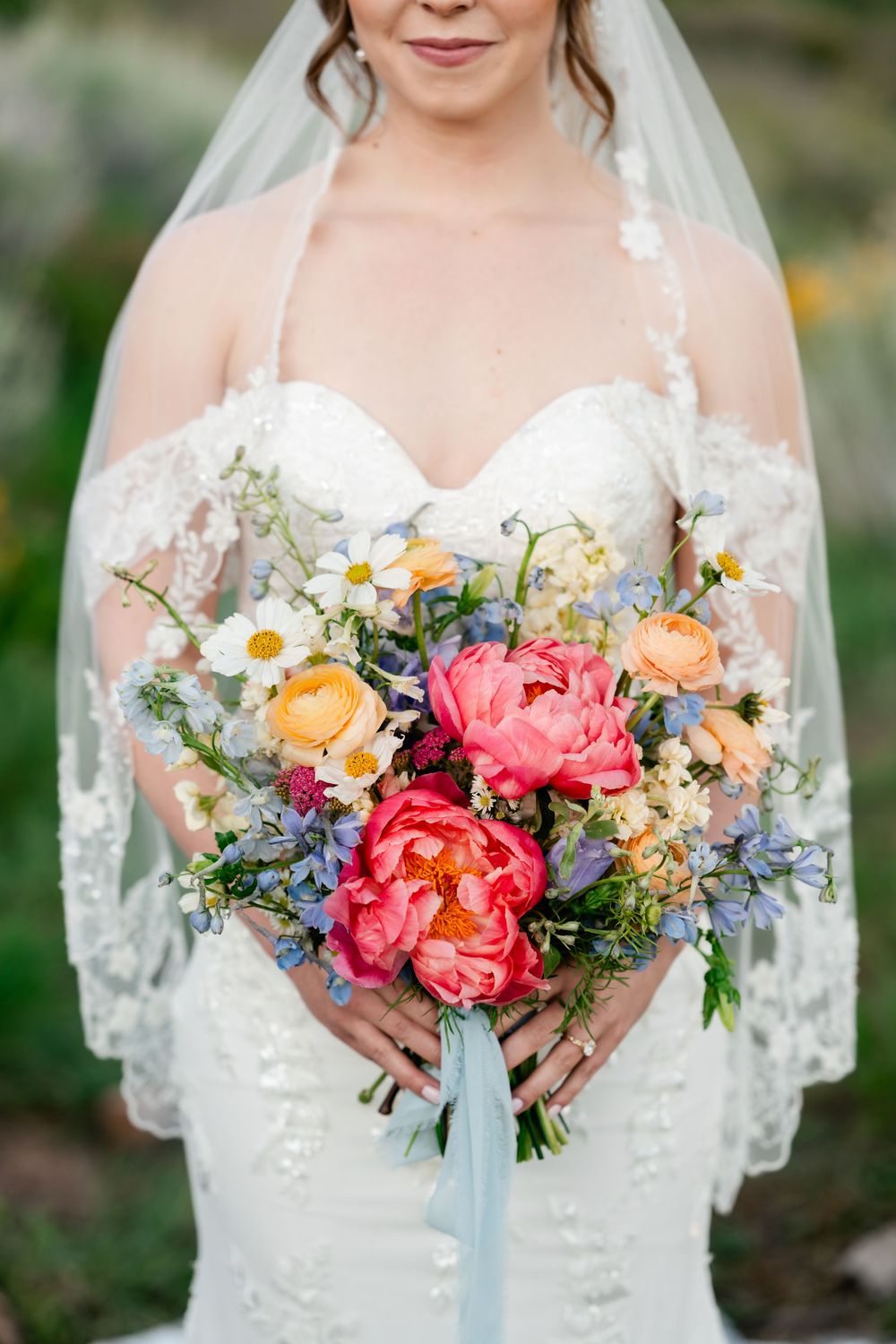 Wedding bouquet close-up shots showing vibrant coral peonies, daisies and blue wildflowers with flowing silk ribbons.