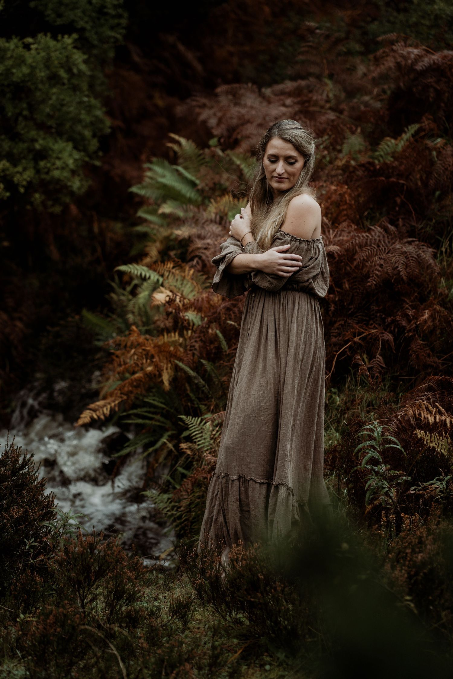 Person in flowing black dress poses dramatically beside waterfall in moody nature setting.