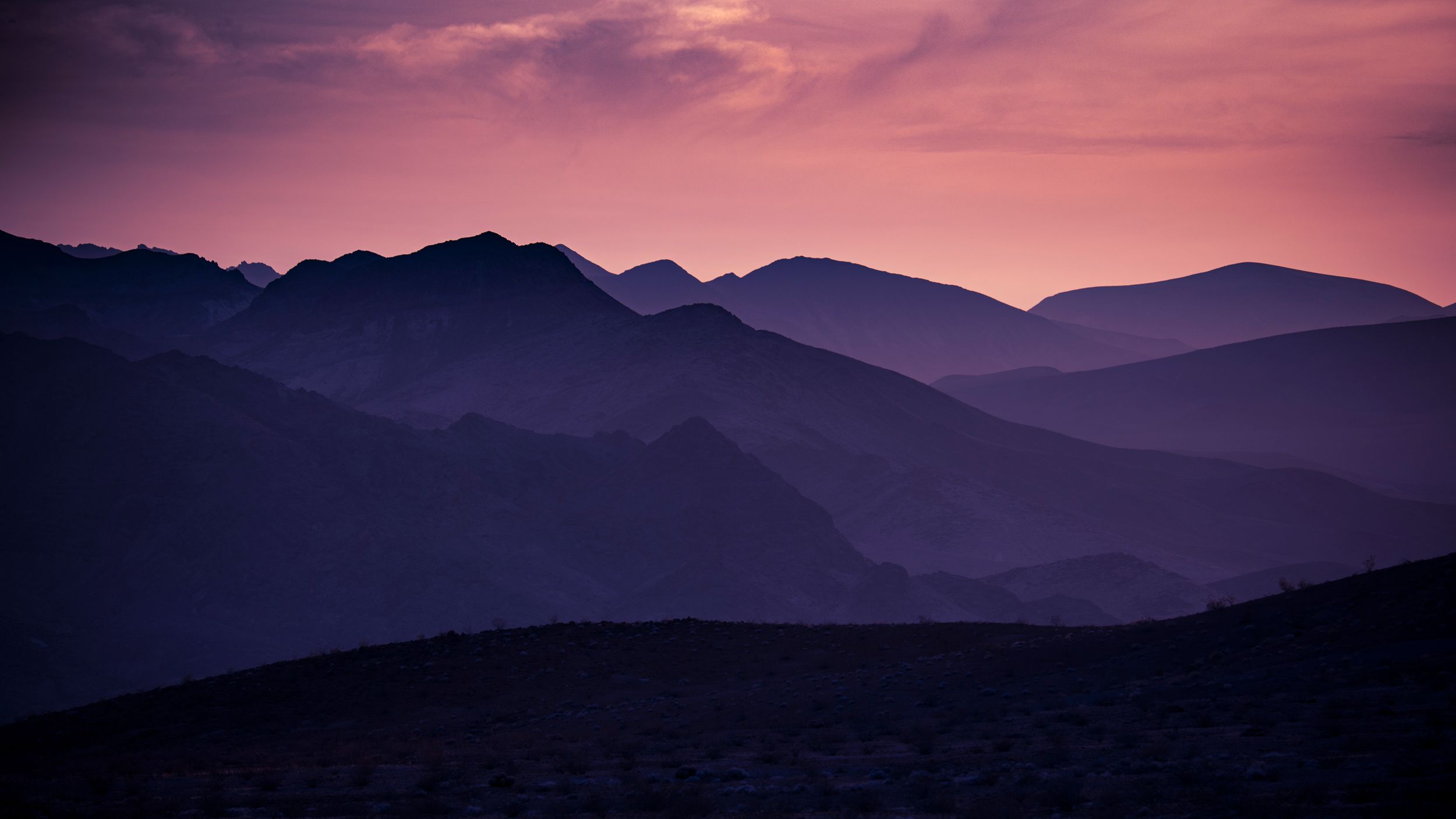 Layered mountain silhouettes fade into purple haze at dusk under a pink and violet sunset sky.