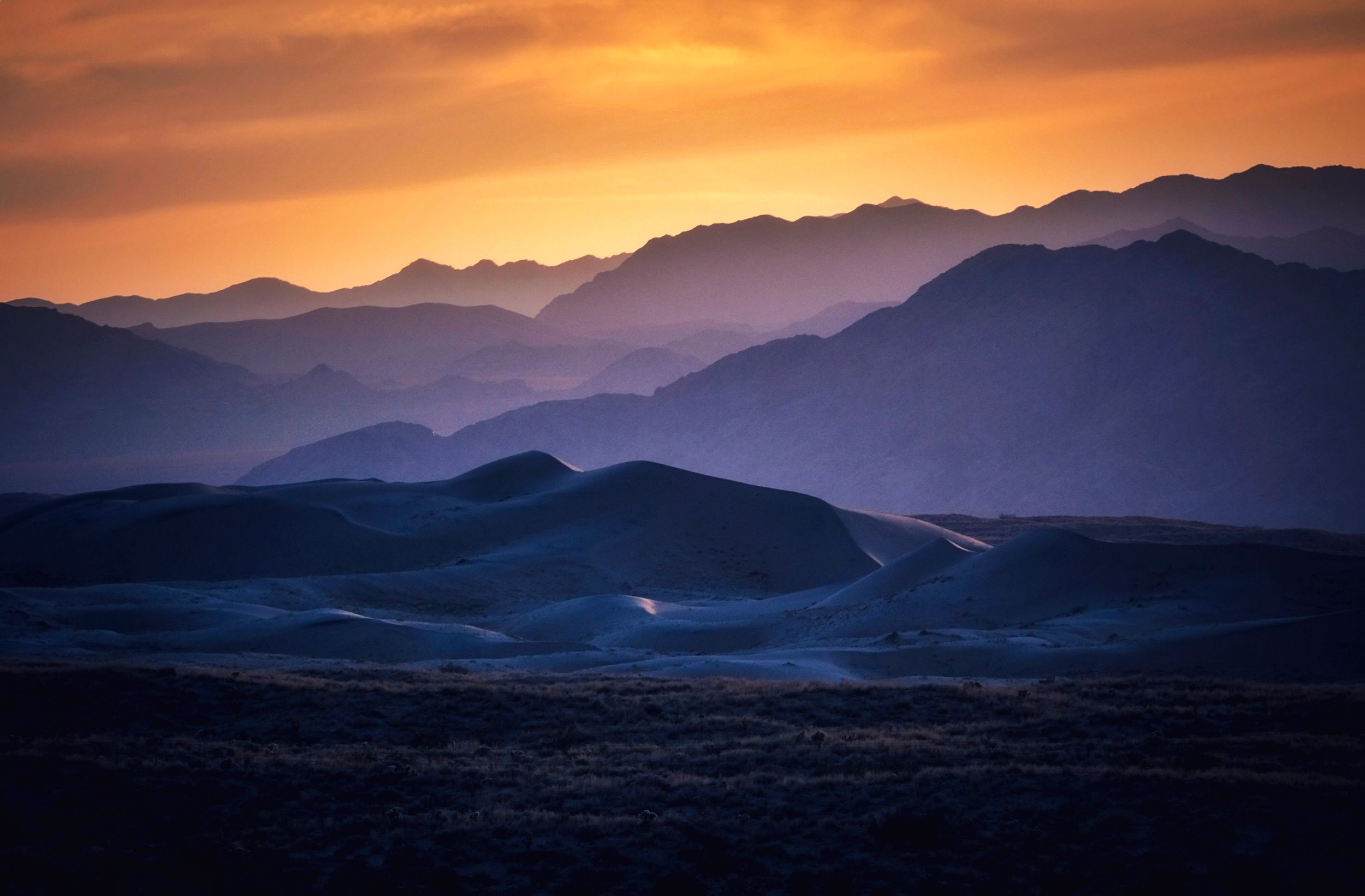 Golden sunset casts warm light over blue sand dunes with layered mountain silhouettes in the background.