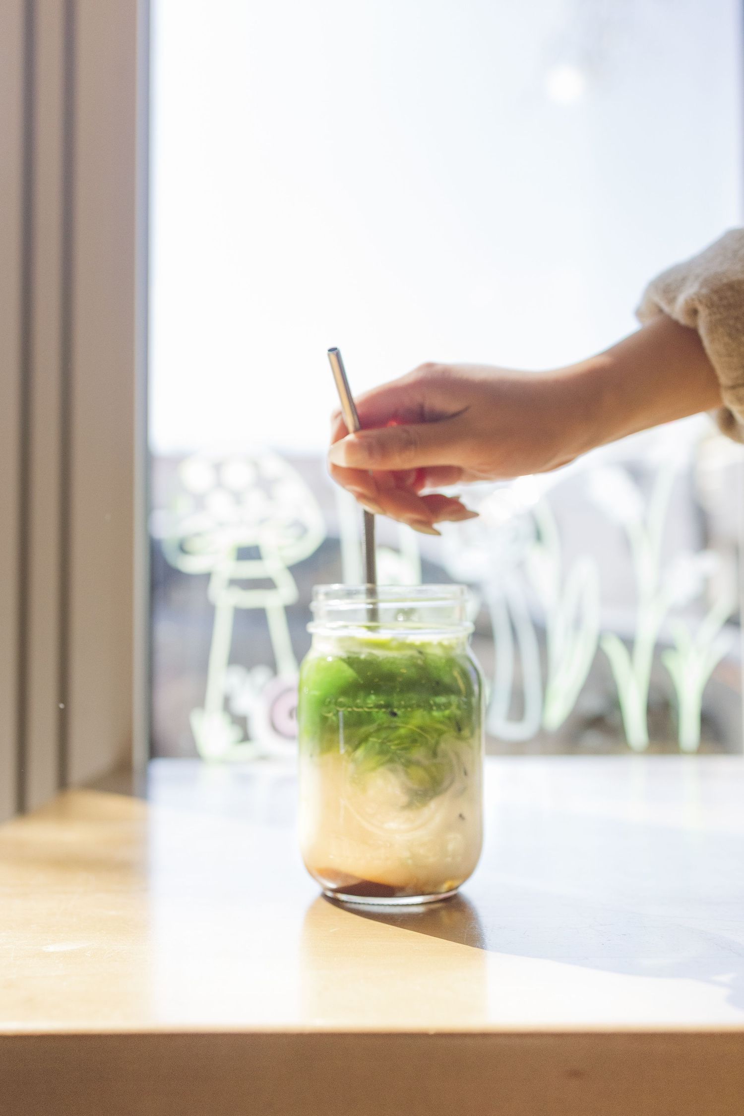 Hands preparing matcha green tea drinks on a wooden table near a window.