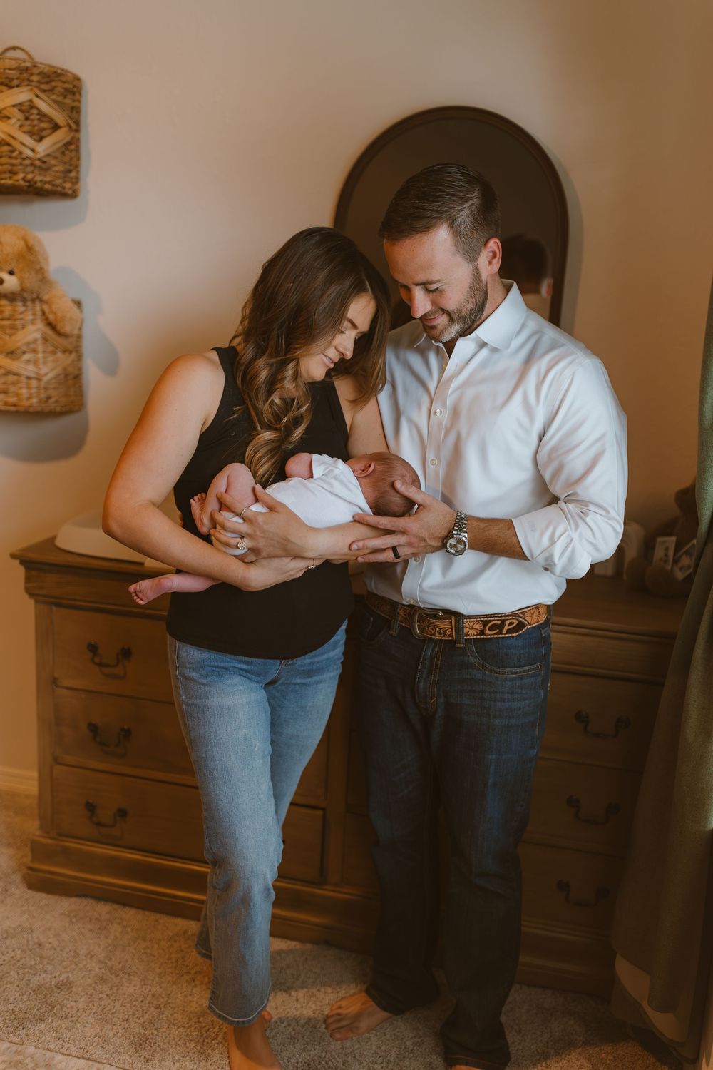 Couple in casual attire tenderly holding and gazing at their newborn baby while standing beside wooden furniture.