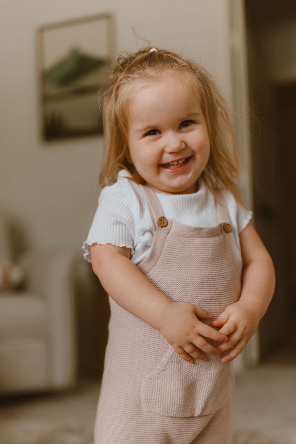 Smiling toddler wearing white t-shirt and beige overalls stands indoors in warm lighting at home.