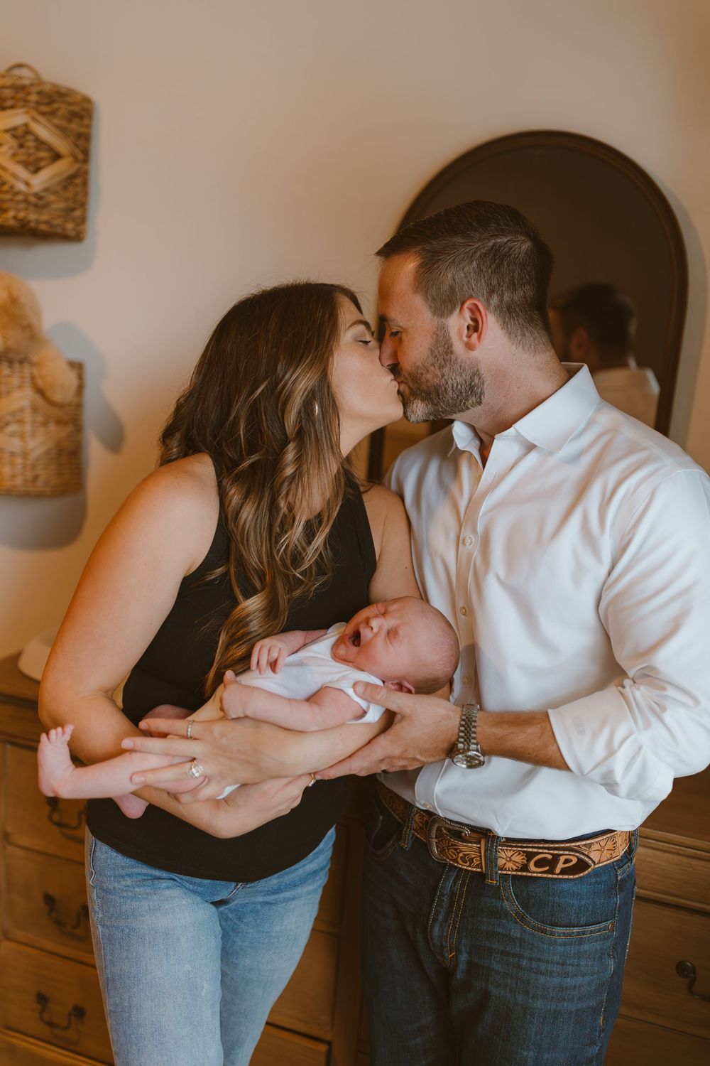 A loving couple share a tender kiss while holding their sleeping newborn baby in a cozy home setting.