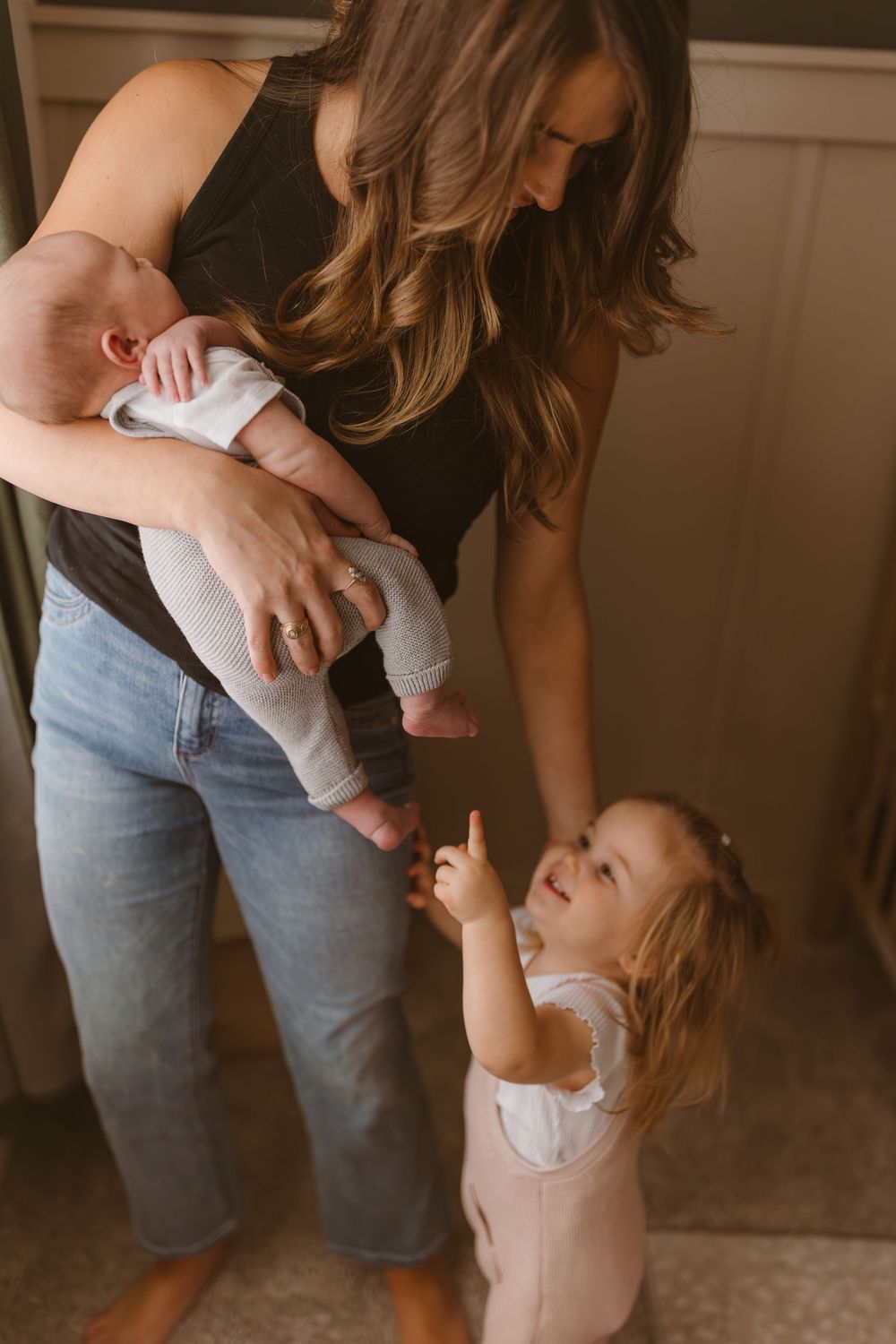 A parent holds a baby while a toddler in a white dress reaches up adoringly in a tender family moment.